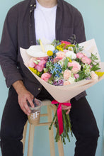 Hombre agarrando con la mano un ramo de flores variado en tamaño, tono y presentación personalizables. Composición floral única seleccionada por nuestras floristas con las mejores flores y fillers disponibles. Foto de ejemplo: Ramo Variado Grande. Vaso de café en la otra mano